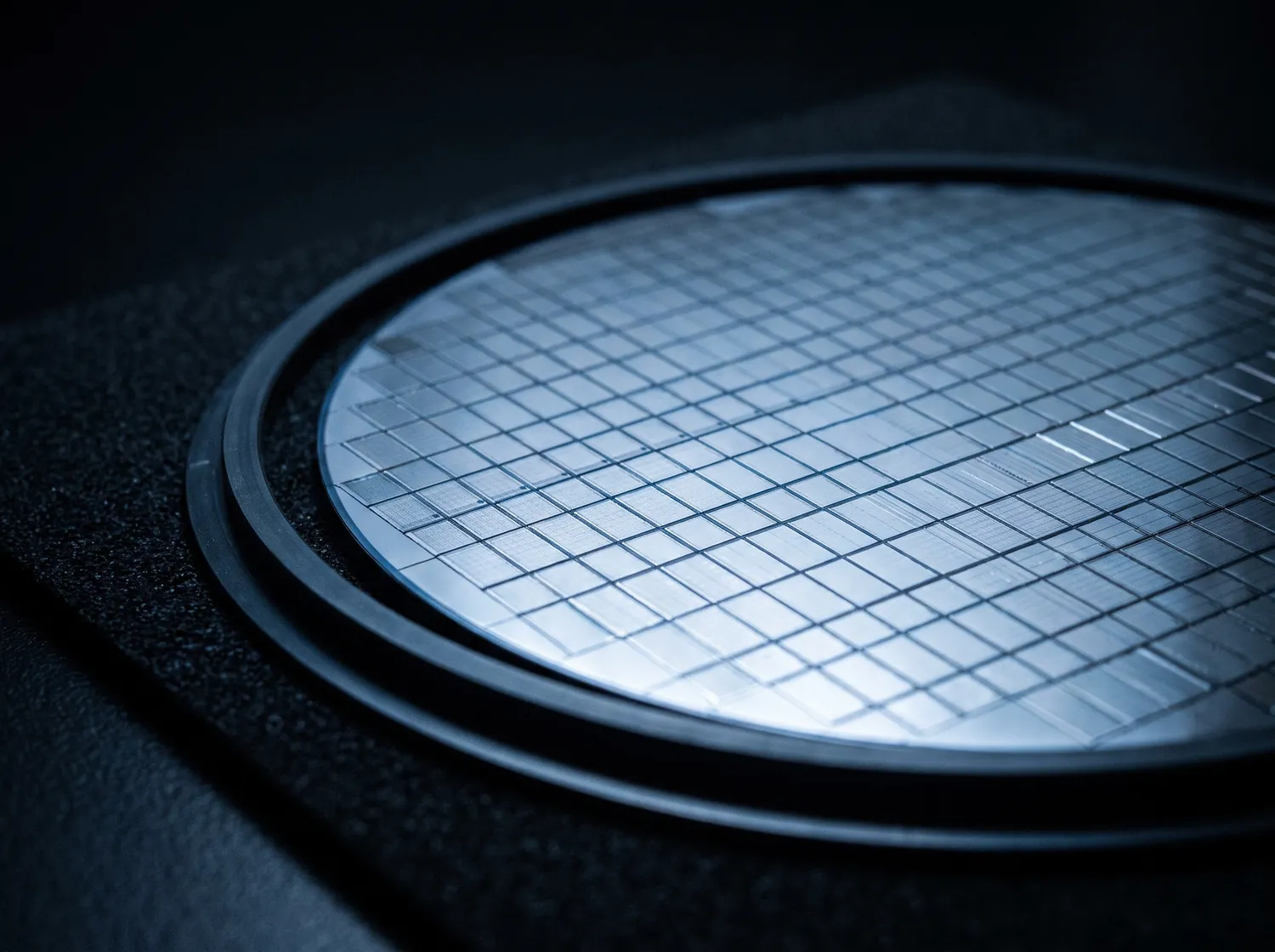 A silicon wafer on a dark bench, its grid of dies catching a cold sheen